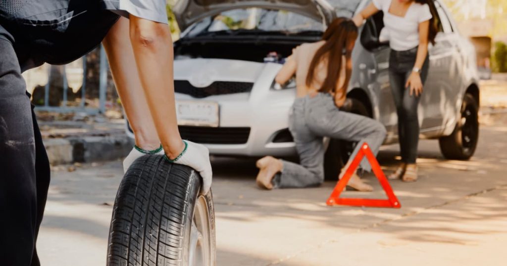 Une voiture avec un pneu crevé est arrêtée et un dépanneur arrive avec un nouveau pneu.