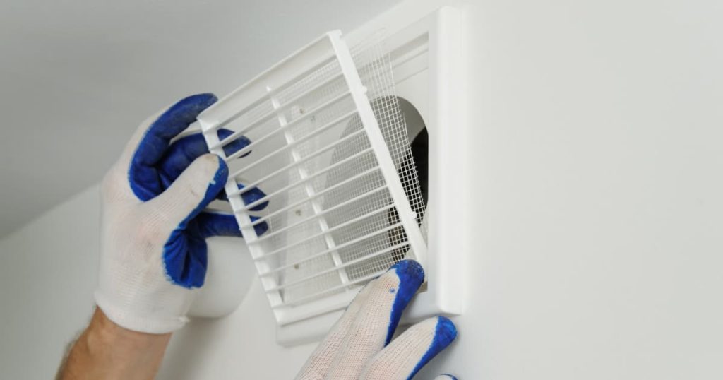 Main ganté entrain de replacer la grille d’un système de ventilation sur un mur blanc