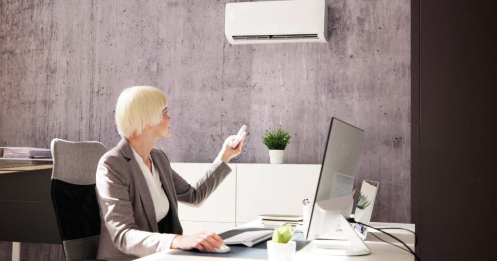 Femme blonde qui manipule son climatiseur grâce à une télécommande en étant assise à son bureau.