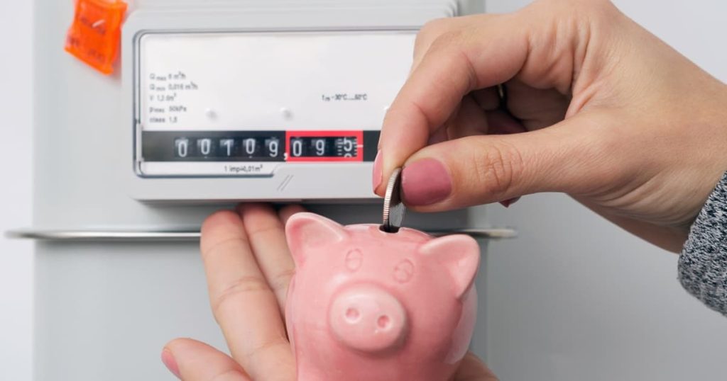 Main mettant une pièce de monnaie dans un tirelire cochon devant un compteur électrique