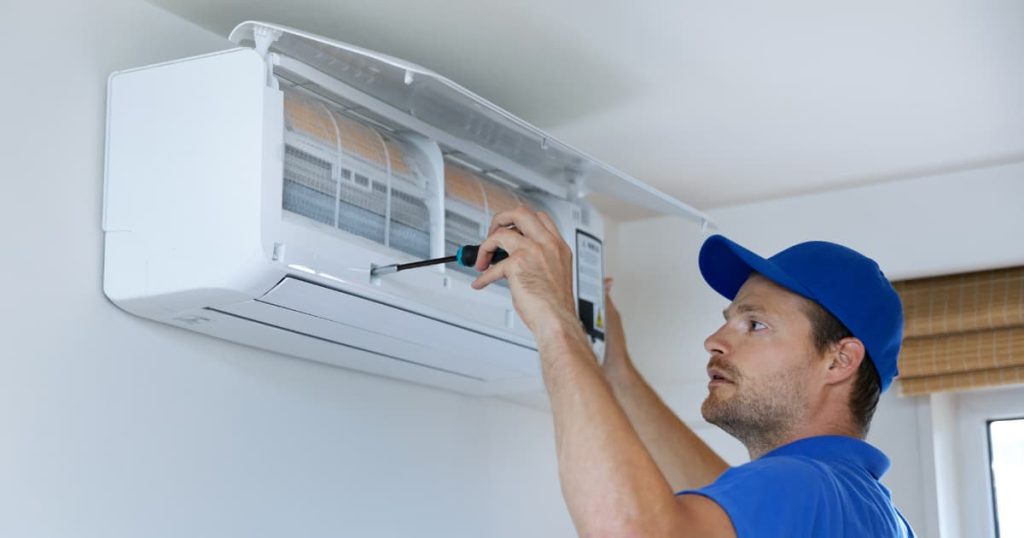 Technicien avec un uniforme bleu et une casquette bleu qui visse un climatiseur fixé en hauteur sur un mur blanc.