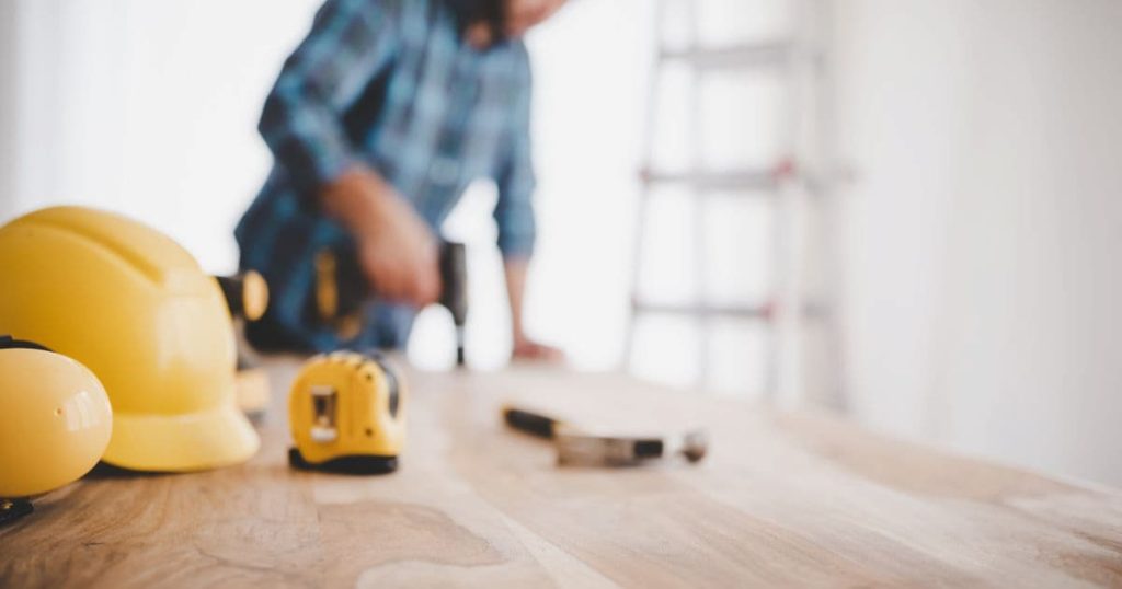 Casques de construction jaunes en perspective sur une table en bois. En arrière plan, un ouvrier en chemise bleu qui avec une visseuse.