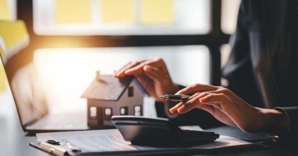 personne qui fait des calculs sur un bureau avec un stylo à la main, une calculatrice, un ordinateur et une petite maison 