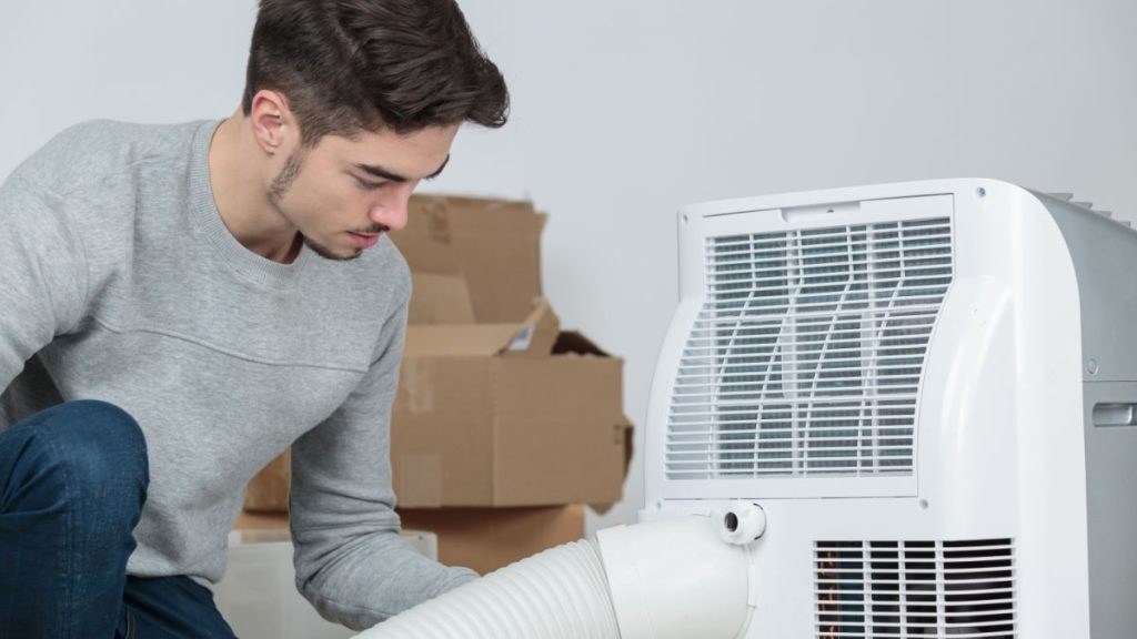 Een man installeert een mobiele airco. Op de achtergrond kartonnen dozen en een witte muur.