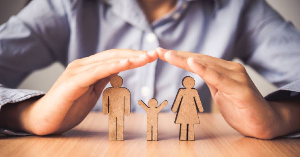 Twee handen vormen een dakje boven drie houten mannetjes: een man, vrouw en kind. Het beeld symboliseert de financiële bescherming van de BA familiale verzekering.