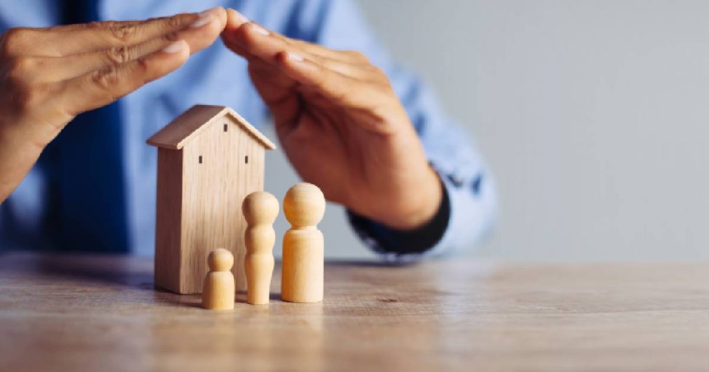 Twee handen boven een houten huisje en 3 houten poppetjes die een gezin voorstellen. Het beeld symboliseert een familiale verzekering (met of zonder franchise).