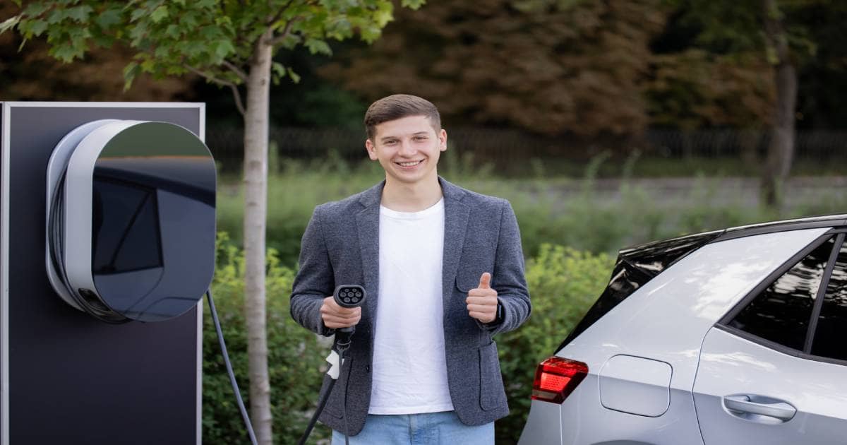 Een blije jongeman laadt thuis zijn elektrische wagen op naast een compacte en stijlvolle laadpaal.