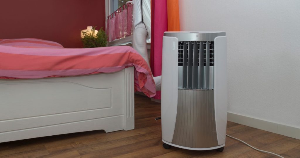 Mobiele airco unit die verplaatst kan worden door de ruimte. Het toestel staat in een slaapkamer met een roze wand en houten vloer. De afvoer van de airco is door het raam naar buiten gehangen. 