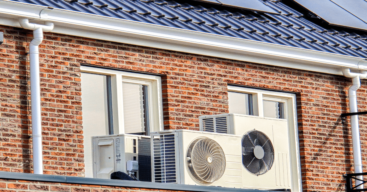 De buitenunit van een split airco systeem op het dak van een woning met bakstenen muur, zwarte dakpannen en zonnepanelen.