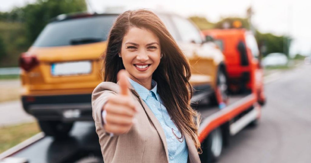 Een dame glimlacht en steekt haar duim op. Op de achtergrond wordt een wagen weggetakeld na een klein ongeval: een kost die ze, dankzij het grondig vergelijken van autoverzekeringen, vergoed krijgt van haar verzekeraar. 