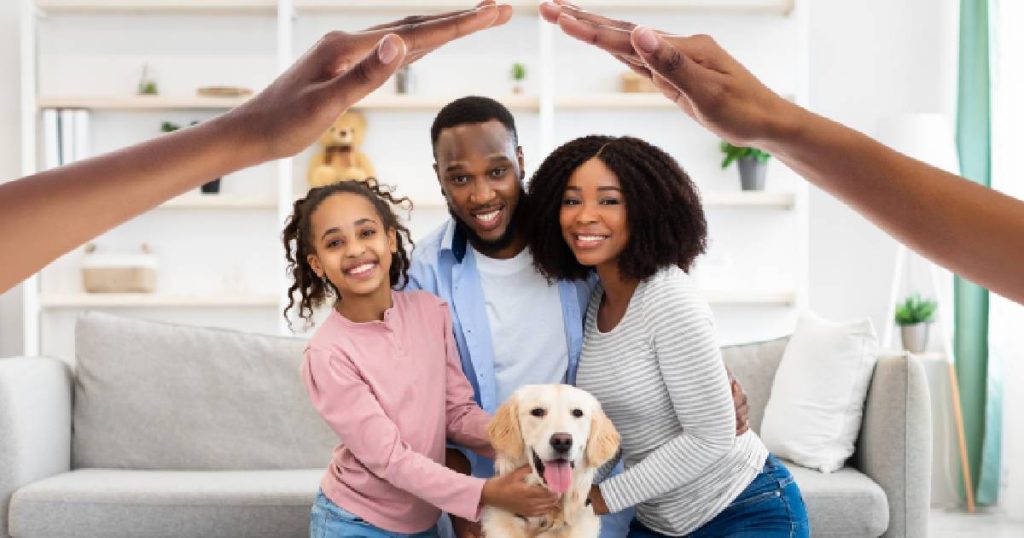 Een familie met vader, moeder, dochter en hond voor een grijze zetel. Boven hun hoofden vormen 2 handen een dak, een symbool voor de financiële bescherming die de familiale verzekering van KBC biedt in ruil voor een eerlijke prijs.