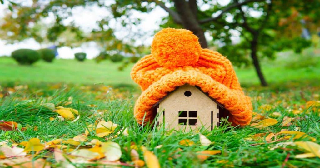 Een houten huisje onder een oranje wollen muts: een symbool voor een goed geïsoleerd gebouw. Het isoleren van je woning kan deels terugbetaald worden dankzij de Mijn VerbouwPremie.