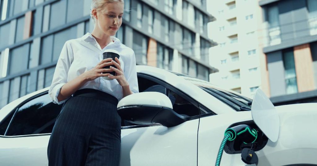 Een dame leunt tegen haar witte elektrische wagen die aan het opladen is via een laadpaal op een parkeerterrein van een appartementsgebouw. De installatie van een laadpaal thuis is nog niet verplicht, maar geldt wel als je bijvoorbeeld een nieuwe woning met 2 of meer parkeerplaatsen bouwt.