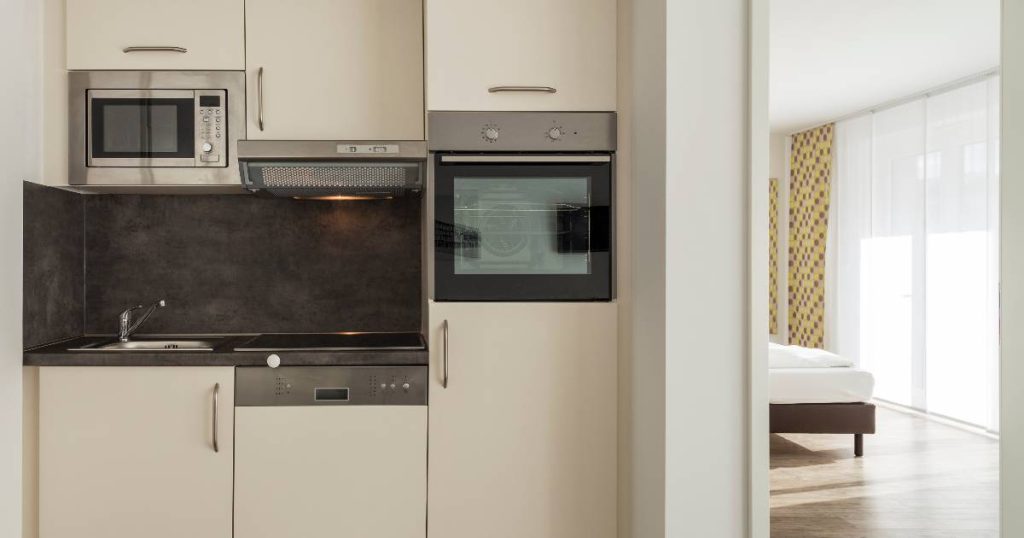 Een kleine modulaire keuken of kitchenette met ingebouwde koelkast, oven en microgolfoven, een spoelblok en een kookblok met kookplaat en afzuigkap. Rechts geeft de keuken uitzicht op een ruime slaapkamer met raam.