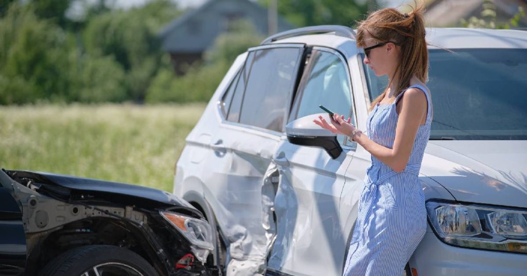 Een jonge dame met rood haar, een blauw met wit gestreepte jurk en een zwarte zonnebril leunt tegen een beschadigde witte auto na een aanrijding. Ze telefoneert met haar autoverzekeraar over de vergoeding van de prijs voor het herstellen van de schade, zoals opgenomen in haar omnium verzekering.