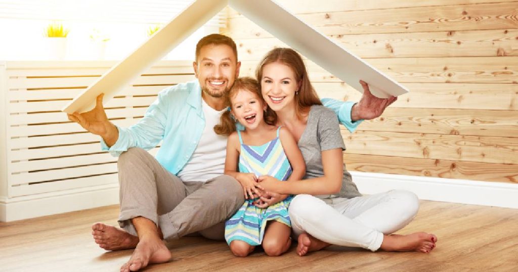 Een man, vrouw en jong meisje zitten samen op een houten vloer. De man houdt een kartonnen bord als dakje boven hun hoofden: een symbool voor de bescherming die een familiale verzekering biedt, in ruil voor een schappelijke prijs.