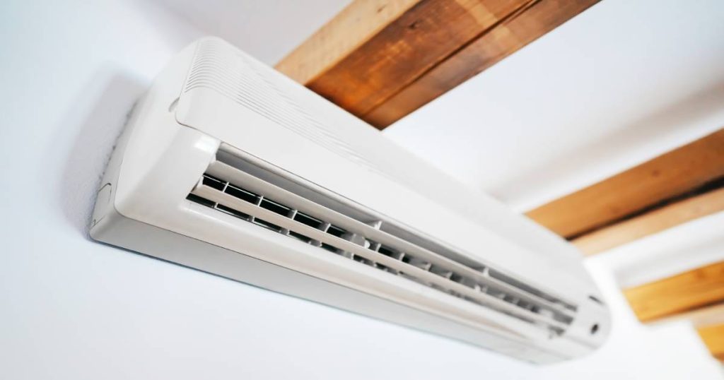 De binnenunit van een split-airco systeem met hoog rendement tegen een witte muur, net onder een wit plafond met zichtbare houten balken.