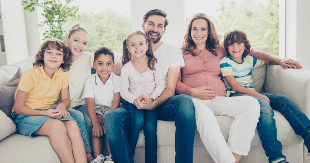 Een groot gezin met een glimlachende vader, moeder, drie zonen en twee dochters op een witte zetel voor een groot raam. Door goed te vergelijken heeft ook dit gezin de juiste familiale verzekering gevonden.