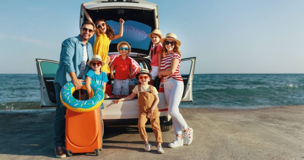 Een moeder, vader en vijf jonge kinderen leunen tegen een witte auto die aan de rand van de zee geparkeerd staat op het strand. De moeder en vier van de kinderen dragen zonnehoedjes, iedereen heeft een zonnebril op. Het gezin is zorgeloos op vakantie nadat het, door goed te vergelijken, de juiste familiale verzekering gevonden heeft.