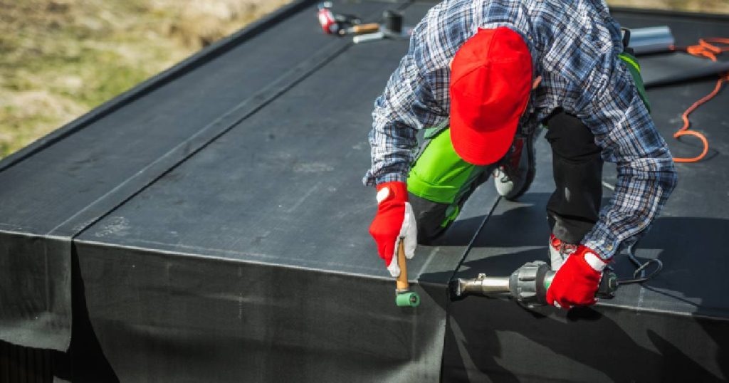 Een man in ruitjeshand met rode handschoenen en pet brengt EPDM dakbedekking aan op het plat dak van een tuinhuis.