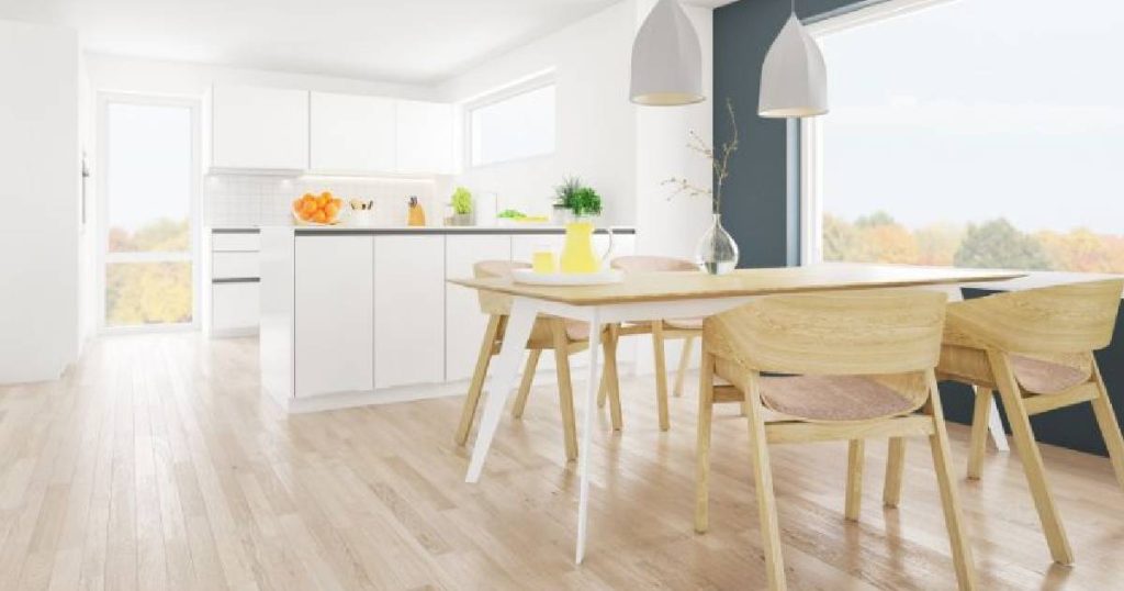 Een moderne Scandinavische keuken met houten vloer en volledig witte keukenkasten. Een eettafel met houten stoelen wordt verlicht door een groot, open raam. Boven de tafel hangen Scandinavische designlampen.