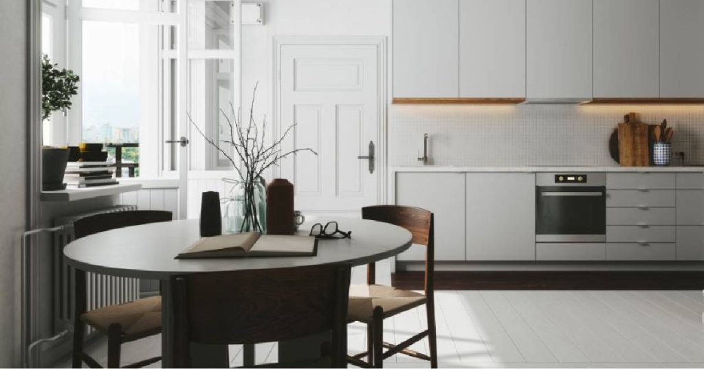 Een sobere, minimalistische Scandinavische keuken met lichtgrijze keukenblokken, een wit stenen werkblad, ingebouwde keukenapparatuur en een ronde, zwarte designtafel met houten stoelen.