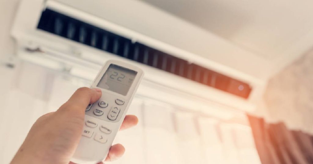 De afstandsbediening van een airco in de hand van een persoon. Op de achtergrond de binnenunit van de airco, die verbonden is met de buitenunit van het vaste split airco systeem.