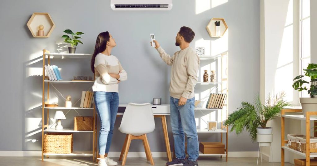 Een koppel staat in een lichte living. De man gebruikt een afstandsbediening om de temperatuur te regelen met de binnenunit van een split airco systeem dat zowel kan koelen als verwarmen.