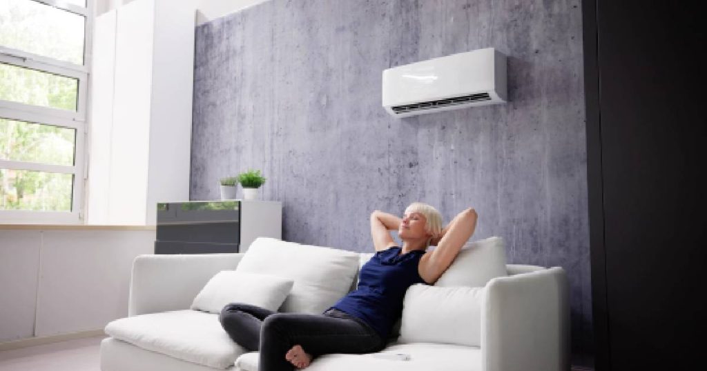 Een dame met lichtblond haar, blauw T-shirt en zwarte broek zit in een witte zetel onder de binnenunit van een airco. De airconditioner hangt tegen een grijze muur.