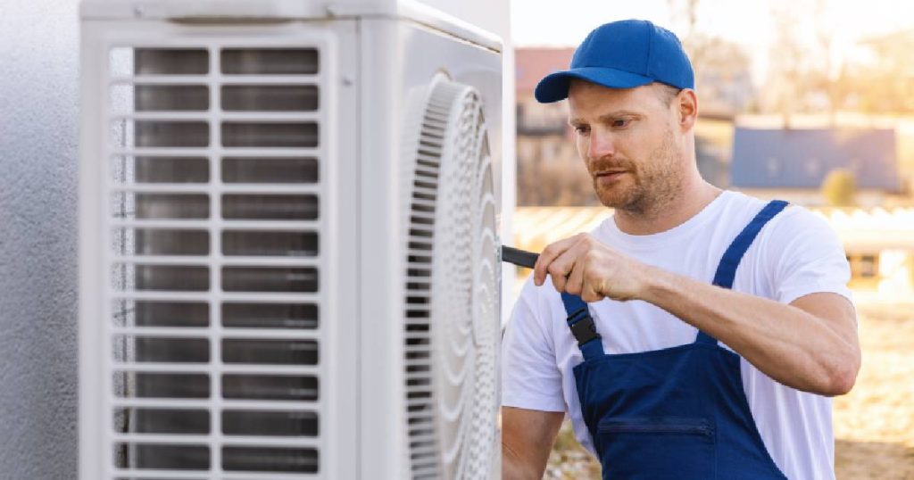 Een aircospecialist installeert de buitenunit van een airco systeem bij klanten die de airco leasen. Hij draagt een wit t-shirt onder een blauwe overall en een blauw petje.