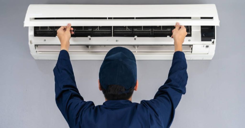 Een installateur in blauwe overall en blauwe pet is in de weer met het plaatsen van een nieuwe witte airco in wandmodel, zoals bepaald op de offerte aan de klant.