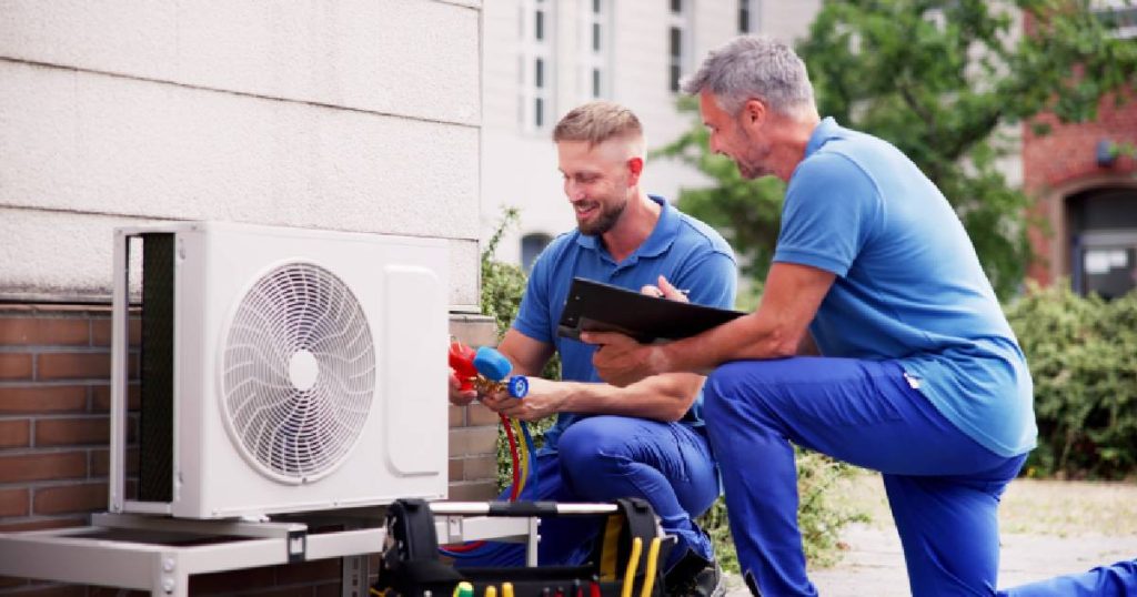 Twee mannen in blauwe broeken en blauwe T-shirts installeren de buitenunit van een nieuwe airco, op de plek die bepaald werd in de offerte;