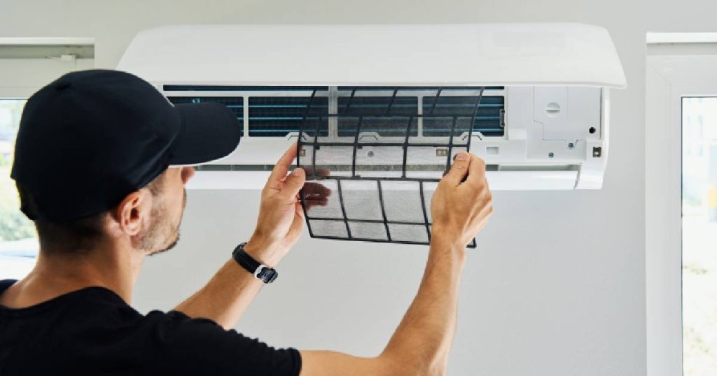 Een installateur vervangt de filter van de binnenunit van een vaste single- of multi-split airco. Hij draagt een zwarte pet, zwart T-shirt en zwart horloge. De airco hangt tegen een muur, tussen twee ramen.