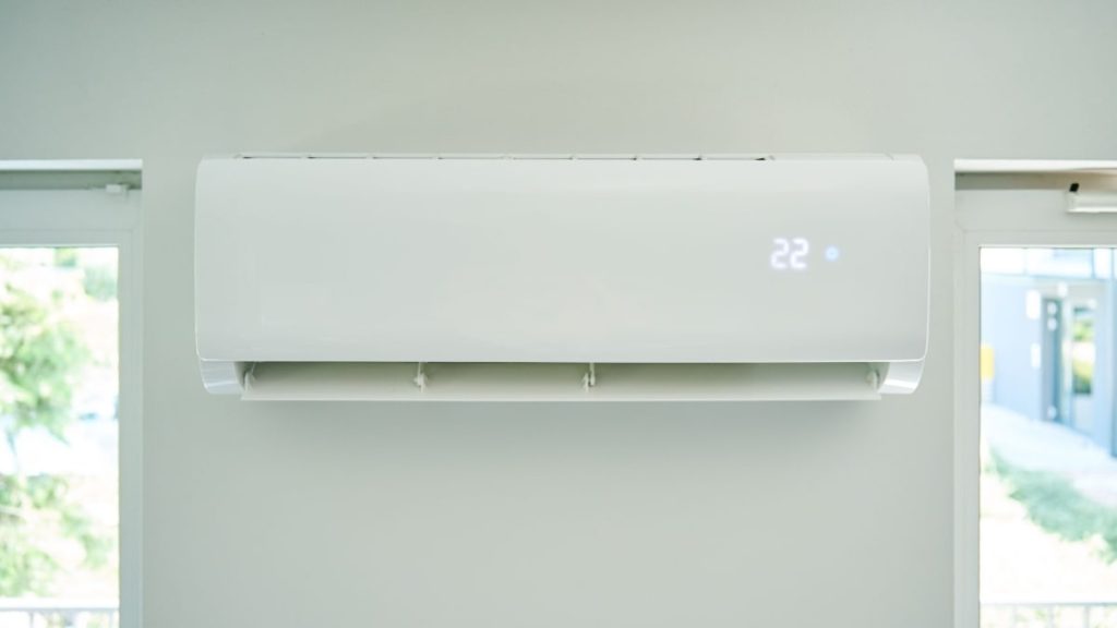 Een witte aircoheater binnenunit tussen 2 ramen, een toestel waarmee je voor een schappelijke prijs je woning kunt koelen en verwarmen. De temperatuur op dit toestel is ingesteld op 22 graden.