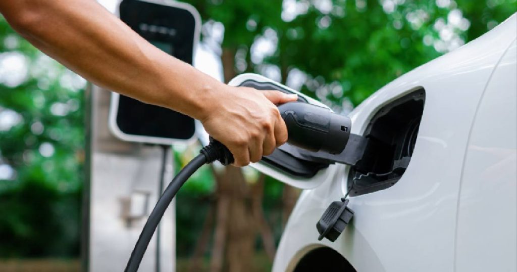 Een hand houdt een laadkabel vast dicht bij de socket van een witte elektrische auto die opgeladen wordt volgens de regels rond thuis opladen. 