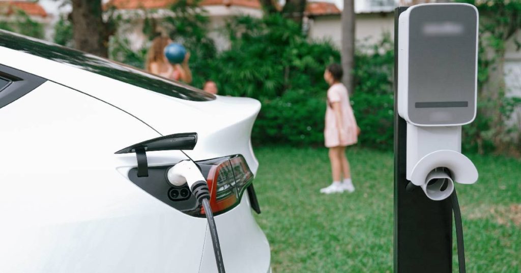 Een witte elektrische auto die thuis aan het opladen is aan een laadpaal, conform de regels. Op de achtergrond spelende kinderen in een tuin.