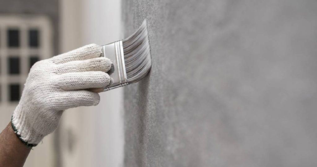 Een man met witte handschoen gebruikt een borstel om een keukenmuur te beschilderen met betonverf en de keuken te voorzien van een betonlook.