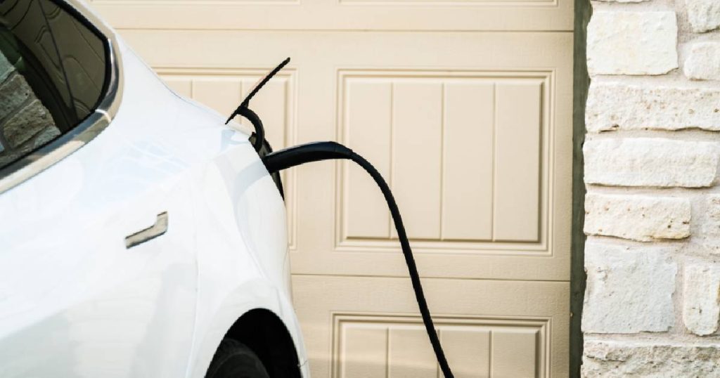 Een witte elektrische wagen voor een lichtgele garagepoort, aangesloten op een slimme laadpaal die bidirectioneel laadt via een zwarte laadkabel.