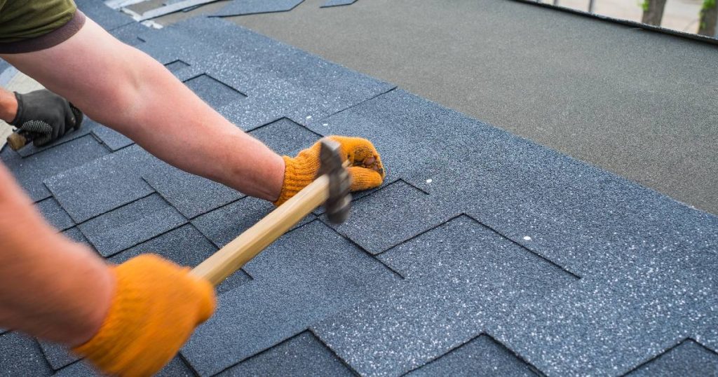 Een dakspecialist gebruikt een hamer en asfaltnagels om bitumen dakshingles op een licht hellend dak te plaatsen.
