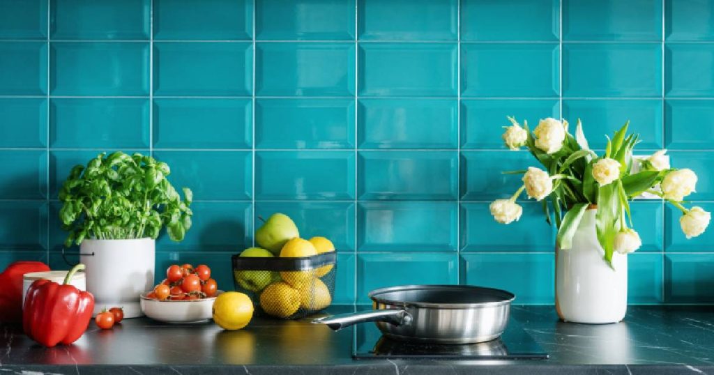 Detailbeeld van een keuken met spatwand in opvallende blauwe tegels en een zwart aanrechtblad. Op het blad staat een steelpan, een witte vaas met gele bloemen, een wit potje met verse basilicum en een verzameling fruit en groenten.