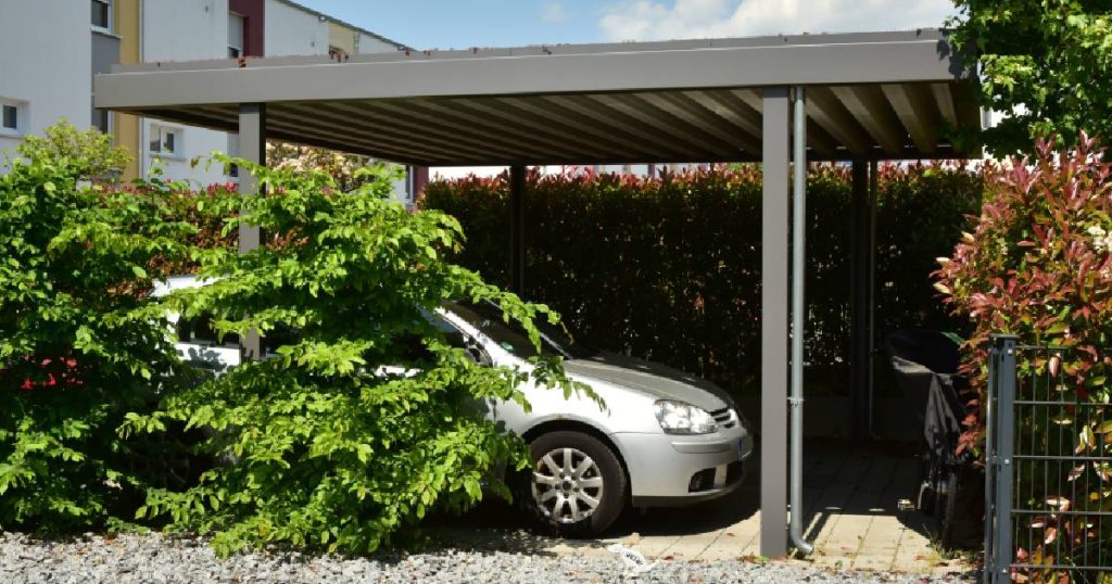 Een moderne, aluminium of stalen carport met houten vloer, omringd door struikgewas. Onder de carport staat een grijze auto. 