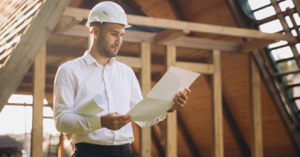 Een architect in wit hemd, witte veiligheidshelm en zwarte broek bekijkt de plannen voor het verhogen van een dak.