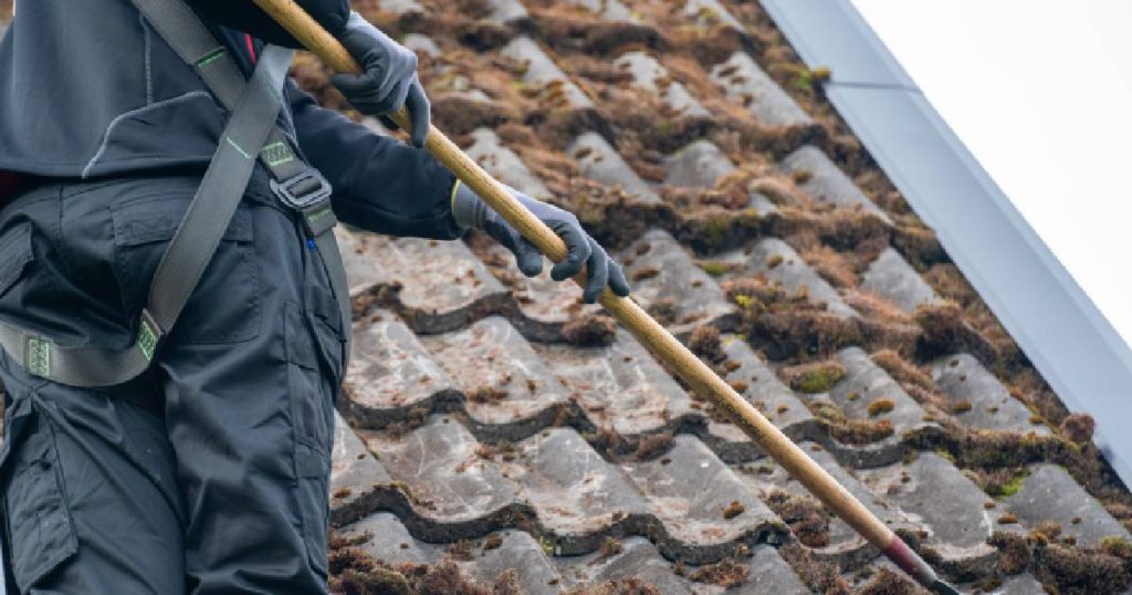 Een specialist gebruikt professioneel gereedschap om mos van een hellend pannendak te verwijderen: belangrijk onderhoud om een daklekkage en schade aan de dakbedekking te voorkomen.