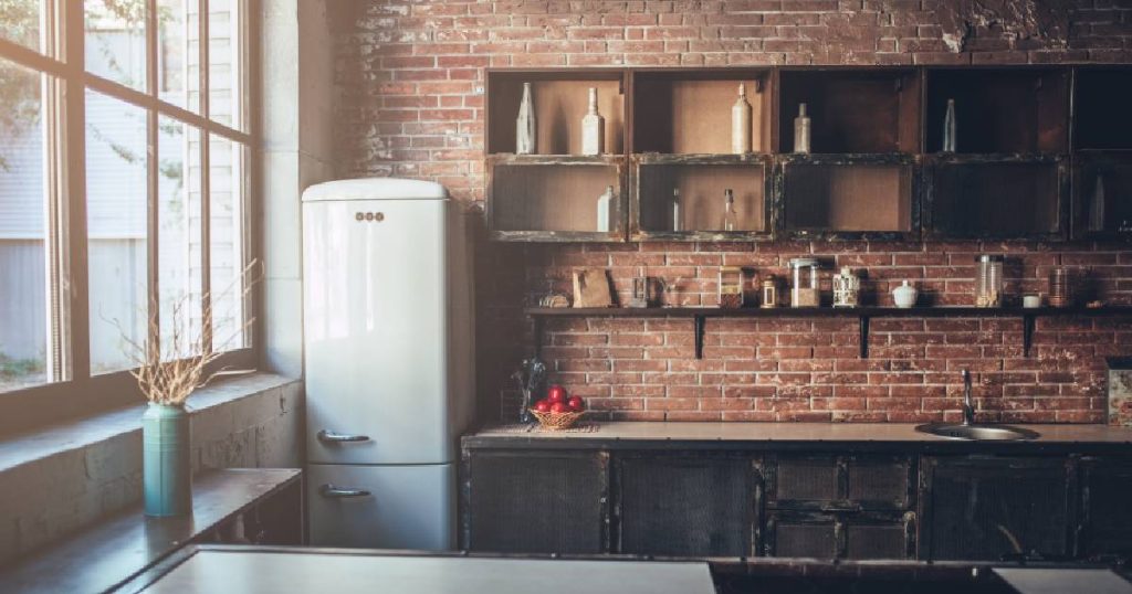 Industriële keuken met donkere open kasten in hout tegen een wand in baksteen. De wandplank wordt ondersteund door donkere stalen L-profielen. De witte frigo is niet ingebouwd en staat los van de rest van de keuken in een hoek.