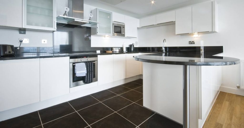 Witte keuken met werkblad in graniet en een donkere vloer die uitgeeft op een laminaatvloer voor de eet-/leefruimte. De dampkap combineert glas en inox. In de kasten boven het aanrecht is verlichting geïntegreerd. Aan de oven hangen 2 handdoeken op.