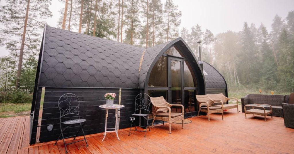 Een kleine moderne woning met gebogen dak en zwarte dakbedekking op een houten ondergrond met terras. In het midden van het klassieke tondak zit een spitsboogdak met grote glazen gevel en glazen deur.