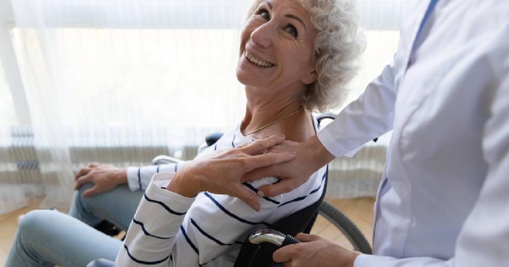 Een dame zit in een rolstoel terwijl ze vriendelijk glimlacht naar de arts die een hand op haar schouder legt tijdens een opname in het ziekenhuis, waarvoor ze een tussenkomst van haar hospitalisatieverzekering geniet.