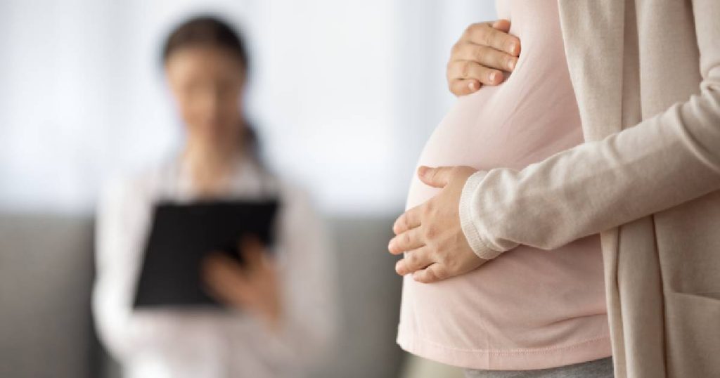 Een zwangere dame in roze t-shirt en lichtroze trui met de handen op de buik, op bezoek bij haar arts, de kosten gedekt door een hospitalisatieverzekering die tussenkomt bij hoge kosten voor zwangerschap en bevalling.