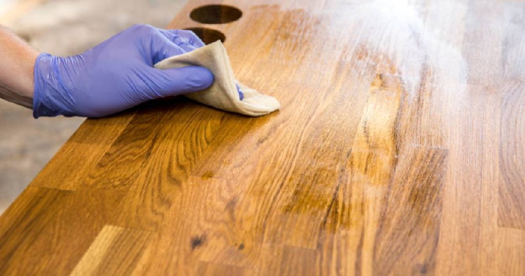 Een hand met blauwe handschoen en een doekje bewerkt een houten keukenblad met olie na het schuren van het keukenwerkblad.