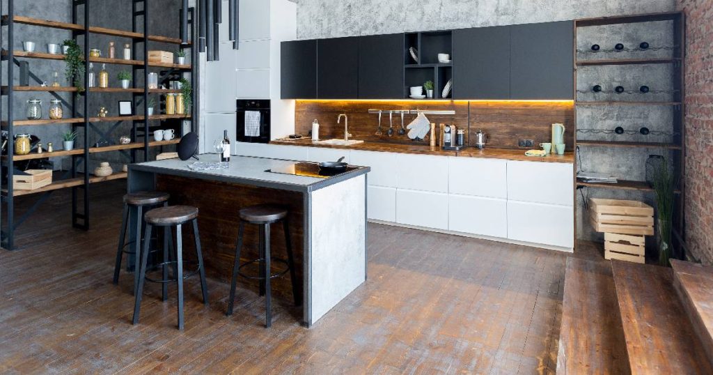 Dezelfde industriële keuken met een strakke, moderne en minimalistische look, dit keer vanuit een andere hoek. De keuken beschikt over een grote kastenwand met spoelbak, een grote, open zwarte stellingkast en een robuust bakstenen kookeiland met kookplaat en toog. De betonnen wand achter de hoge kasten en de stoere hanglampen dragen bij aan het industriële karakter.